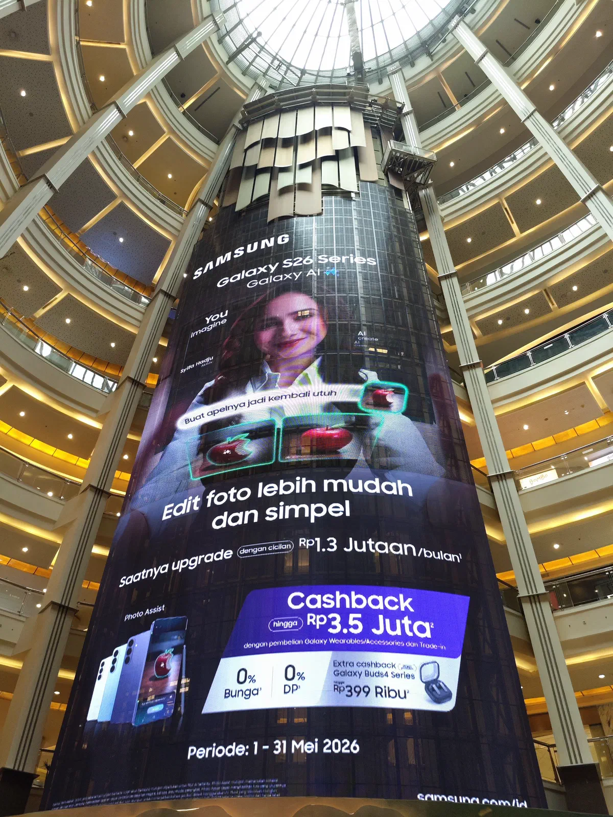 A towering curved LED screen wrapping a column in a multi-storey mall atrium, showing a smartphone campaign